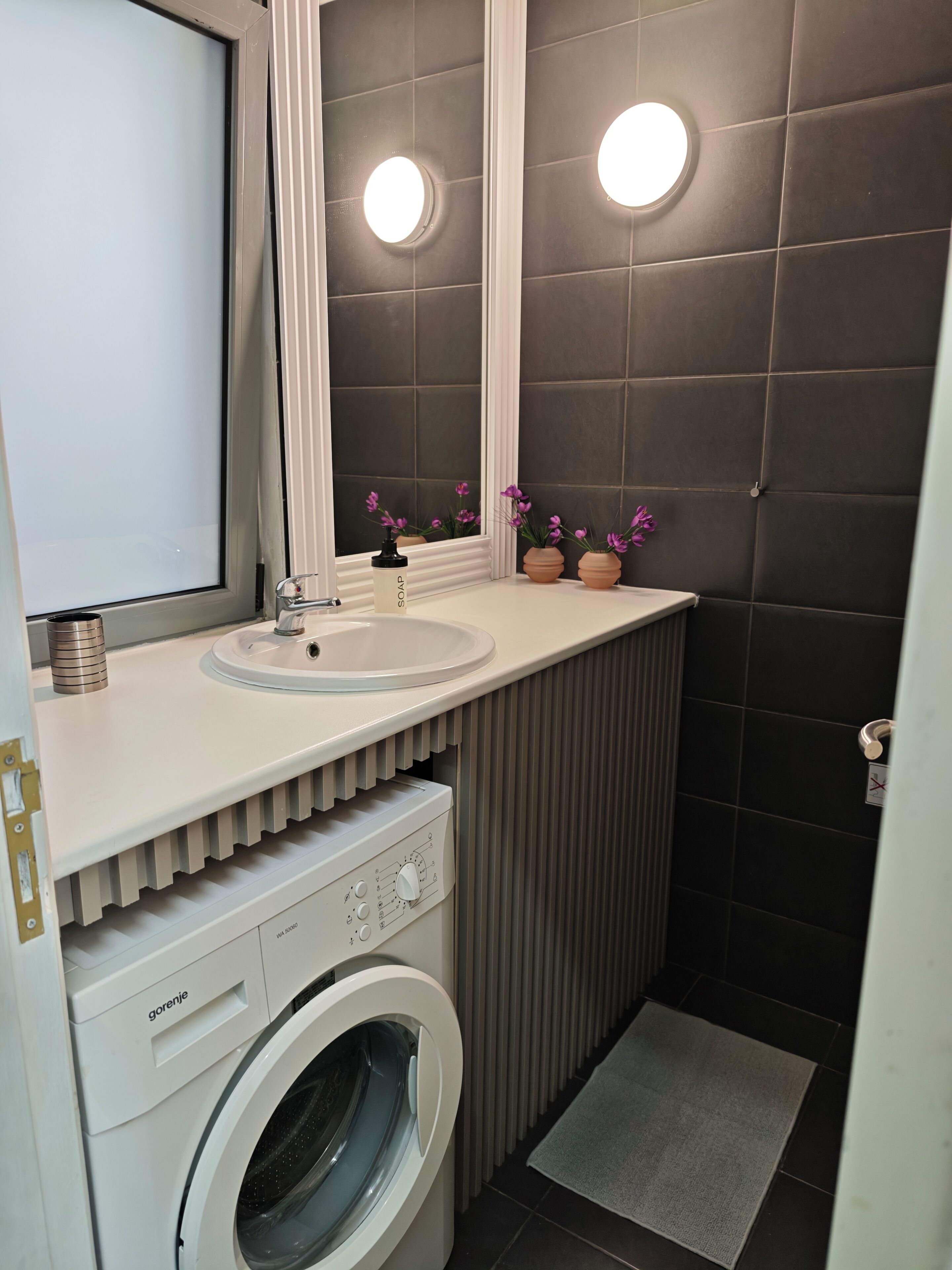 A compact bathroom with a built-in washing machine, a sink, and a mirror.