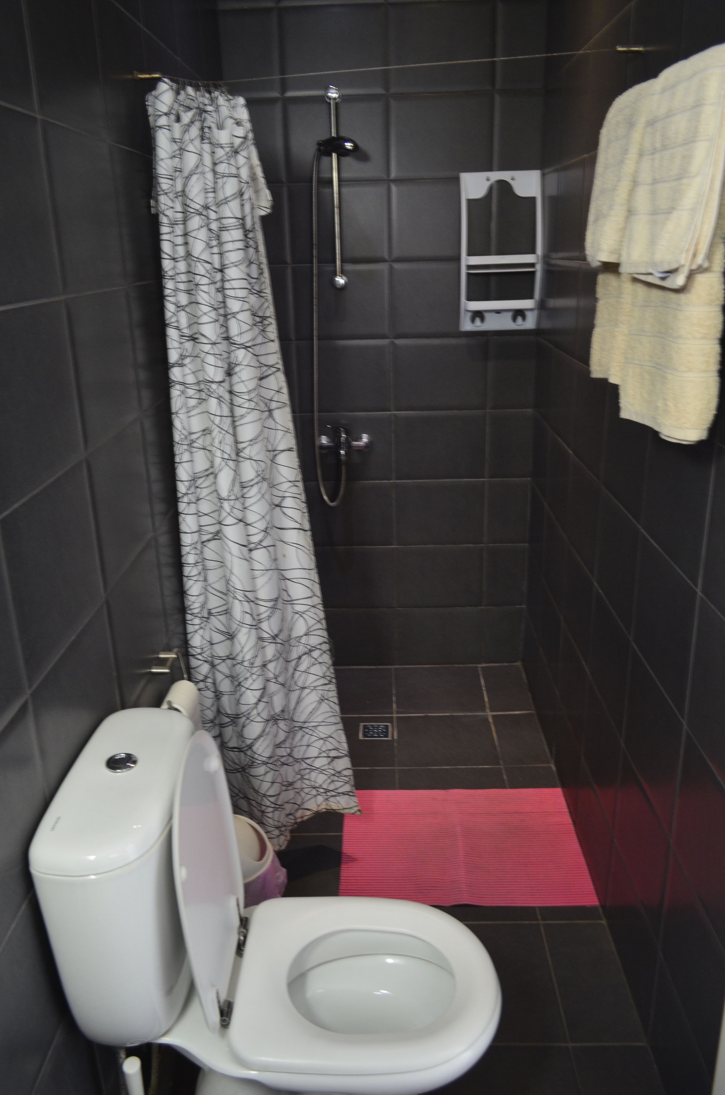 Een kleine, donkere badkamer met een wit toilet, een douche met een decoratieve gordijn en een handdoekrek met een handdoek.