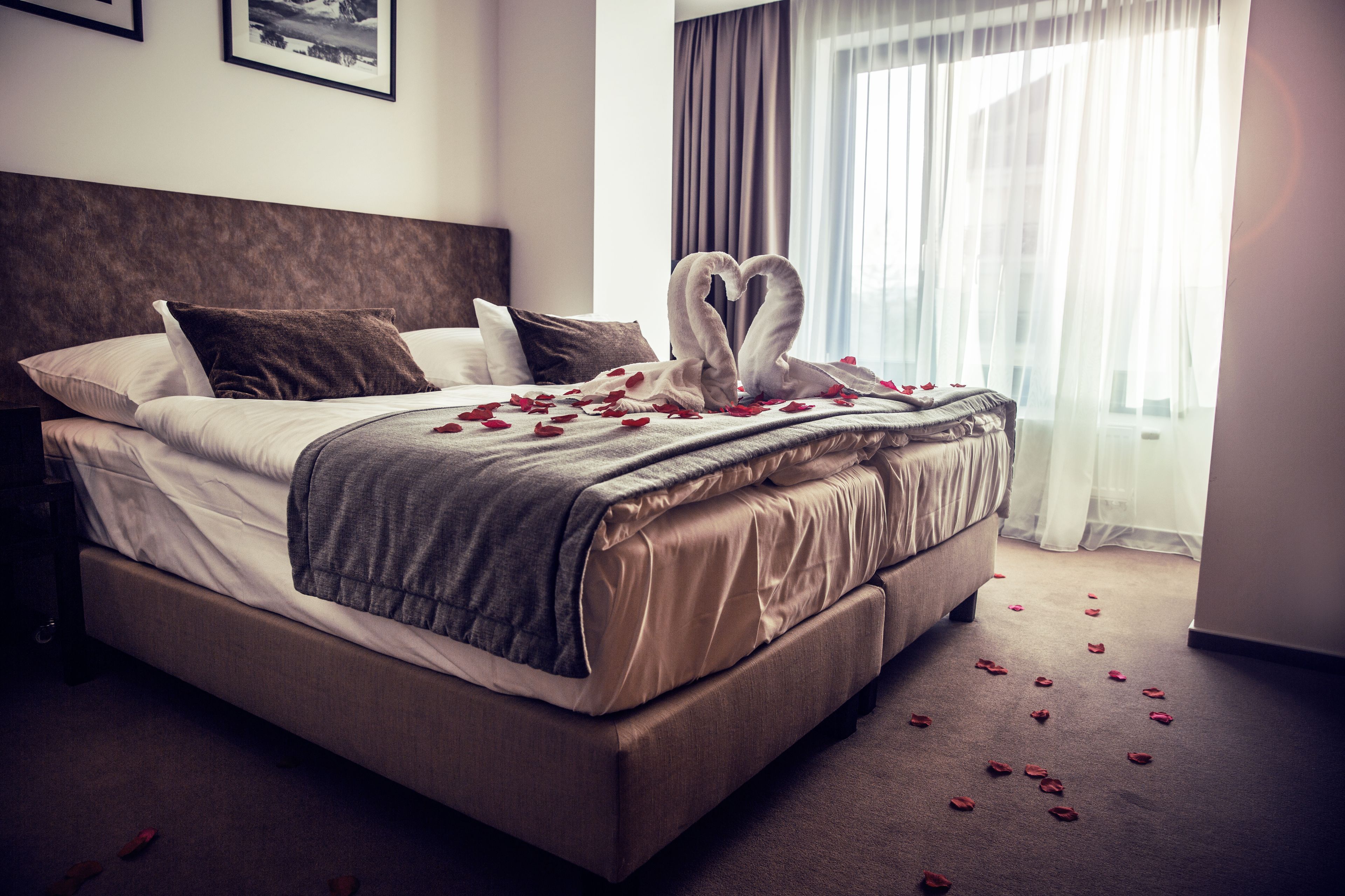 A neatly made bed with a heart-shaped decoration made of rose petals, a brown headboard, and a large window with sheer curtains.