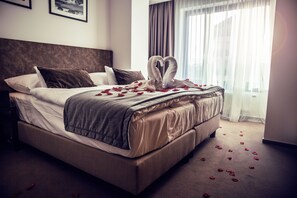 Una cama bien hecha con una decoración en forma de corazón hecha de pétalos de rosa, un cabecero marrón y un ventanal con cortinas de gasa.