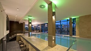 Una piscina interior moderna con barandillas de vidrio, paredes de piedra y vistas a la ciudad al anochecer.