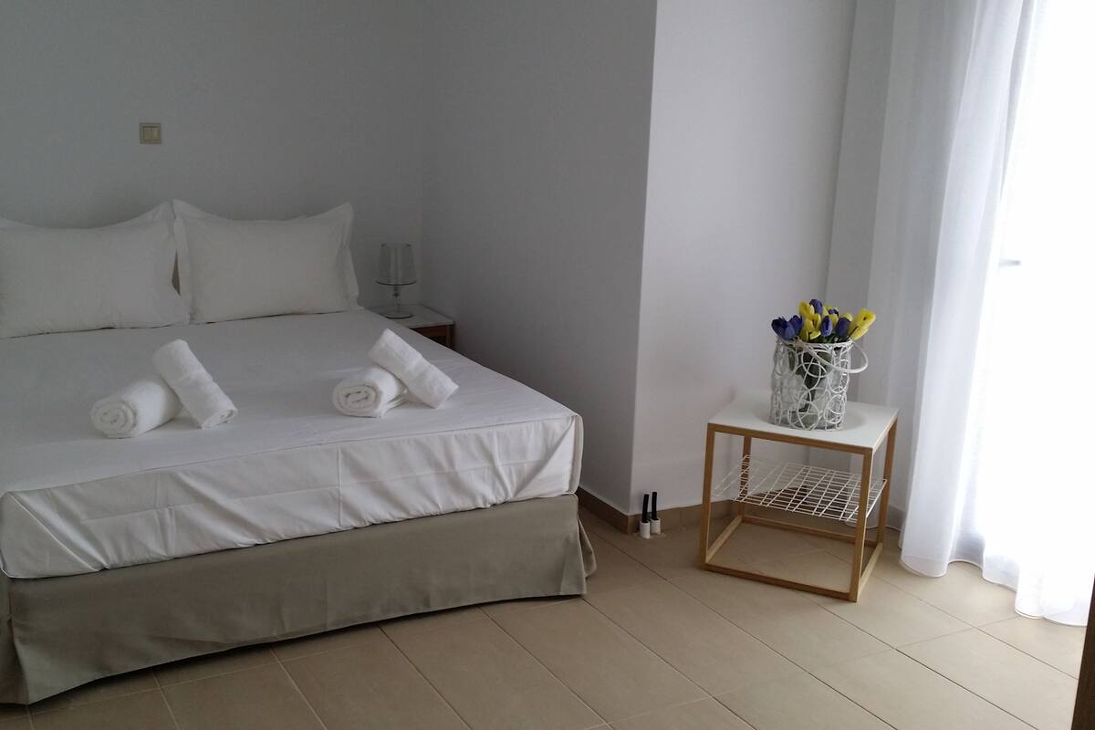 Ein ordentlich bezogenes Bett mit weißen Leinentüchern und zwei aufgerollten Handtüchern, ein Nachttisch mit Lampe sowie ein kleiner Tisch mit Blumen in einer Vase.