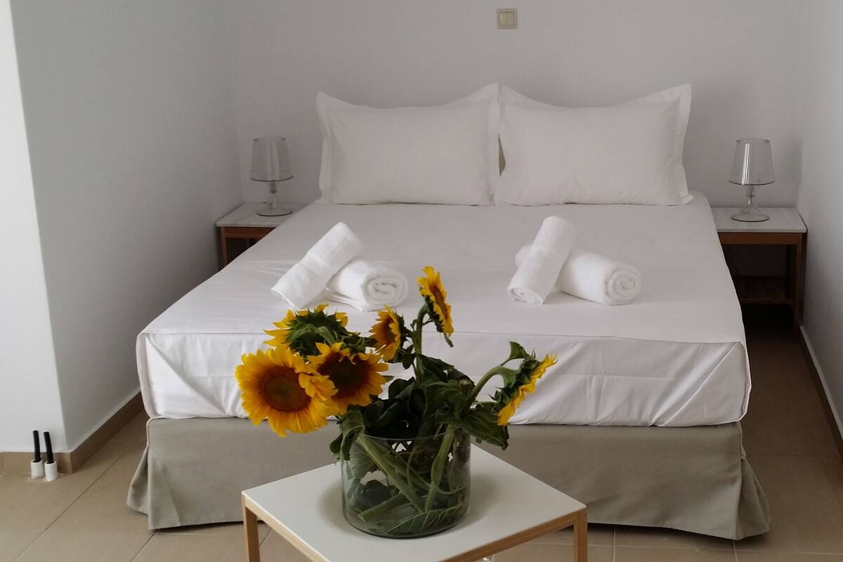 Ein ordentlich bezogenes Bett mit weißen Laken, zwei zusammengerollte Handtücher und eine Vase mit Sonnenblumen auf einem Nachttisch.