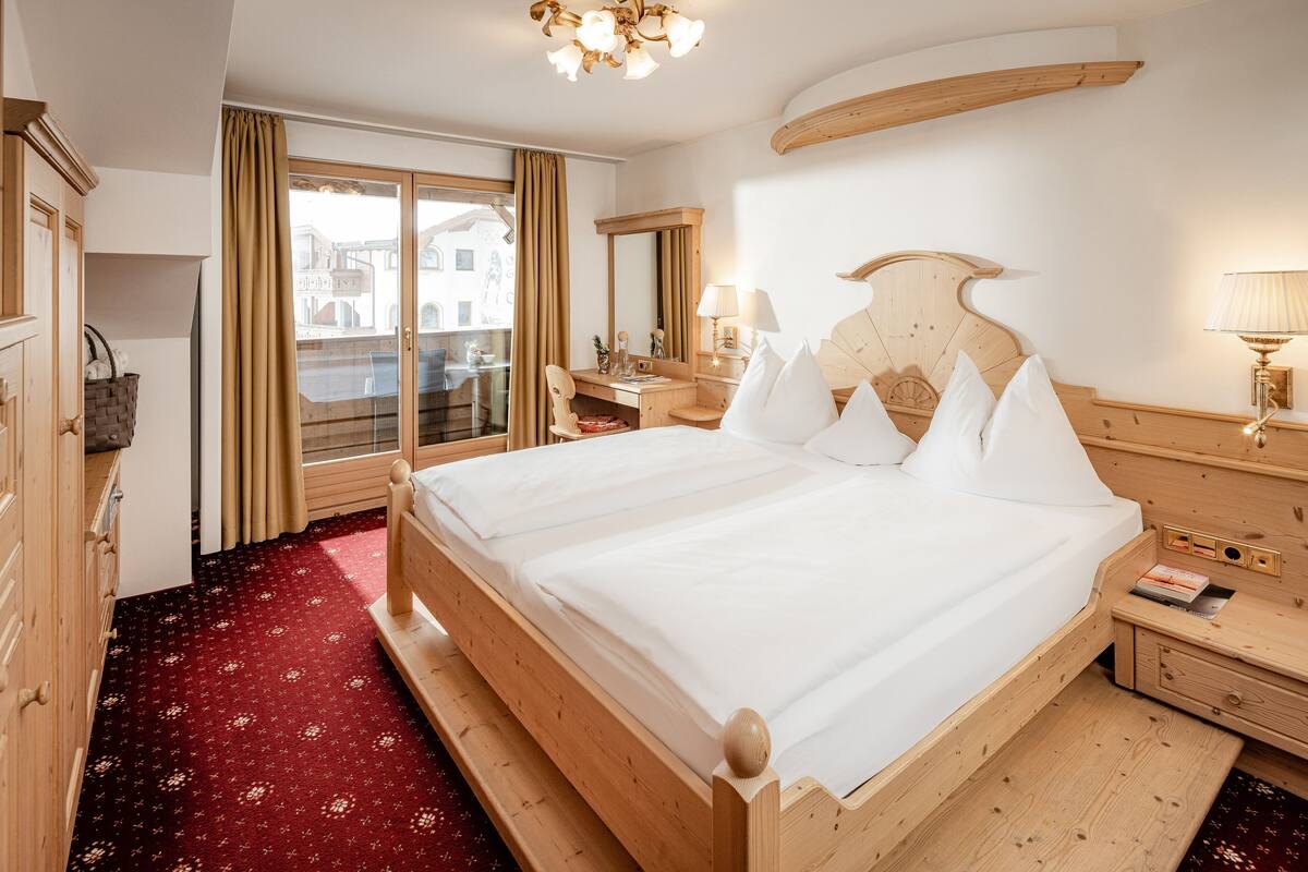 A hotel room with a large bed, wooden headboard, a desk with a chair, a mirror, and a window with curtains.