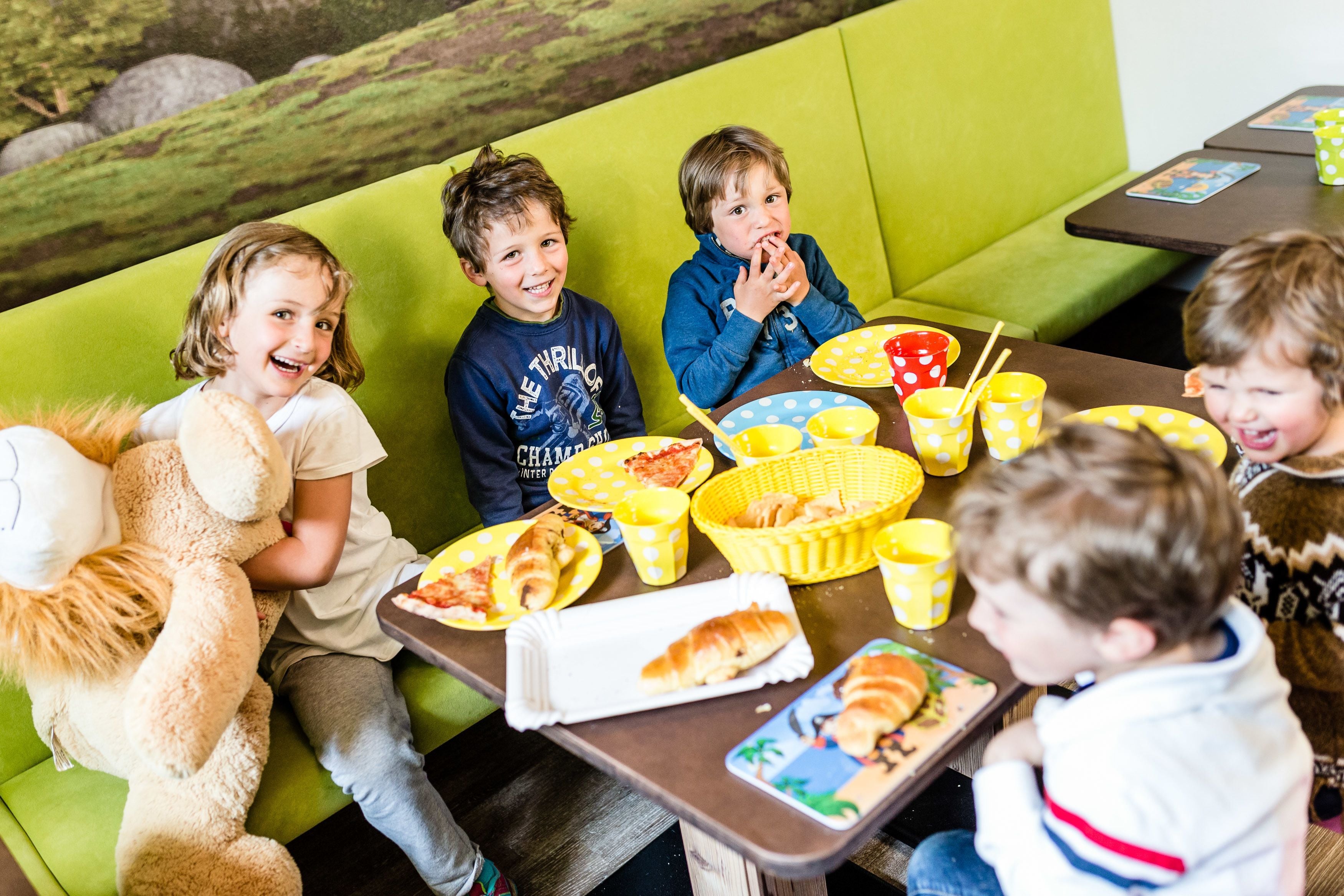 A group of children seated on a green couch at a table with food and drinks.