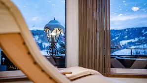 A lounge chair facing a window with a view of a snow-covered mountain and a lit lantern. - Romantic & Family Hotel Gardenia (Selva di Val Gardena)