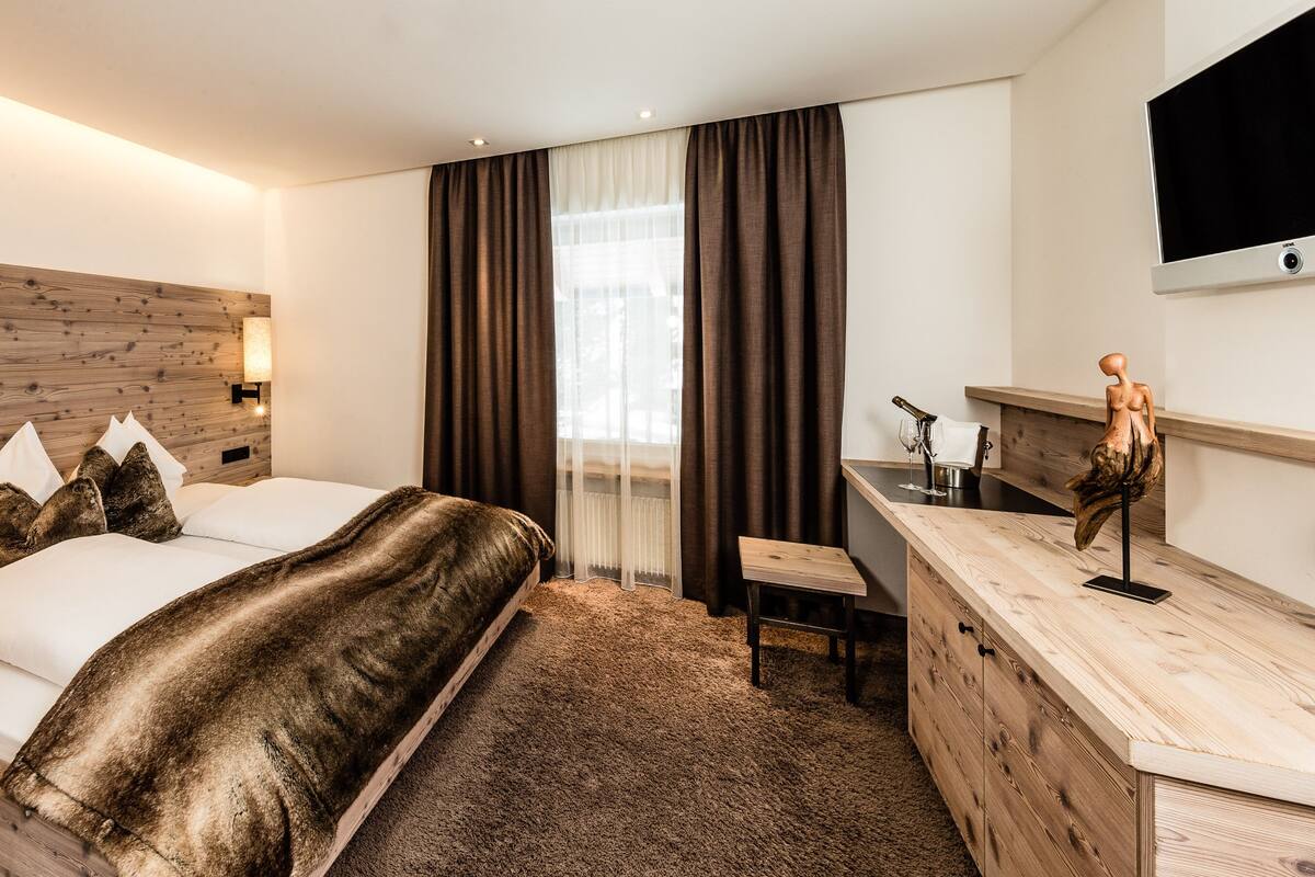 Ein modernes Hotelzimmer mit Holz-Kopfstütze, einem großen Bett mit weißer und brauner Bettwäsche, einem Schreibtisch mit Flachbildfernseher und einem großen Fenster mit Vorhängen.