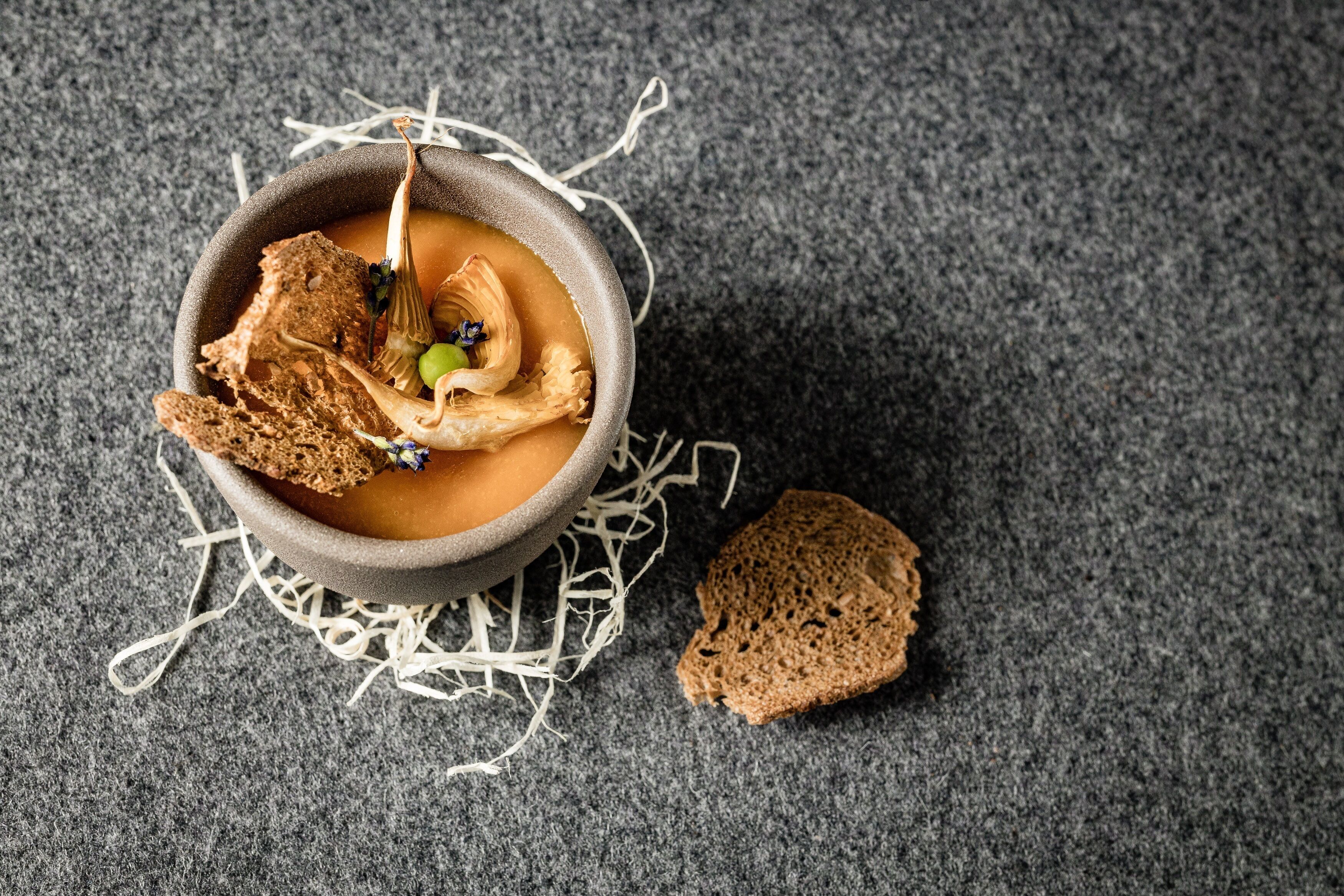 En skål suppe med et stykke brød og en kvist krydderurt på en ru overflade.