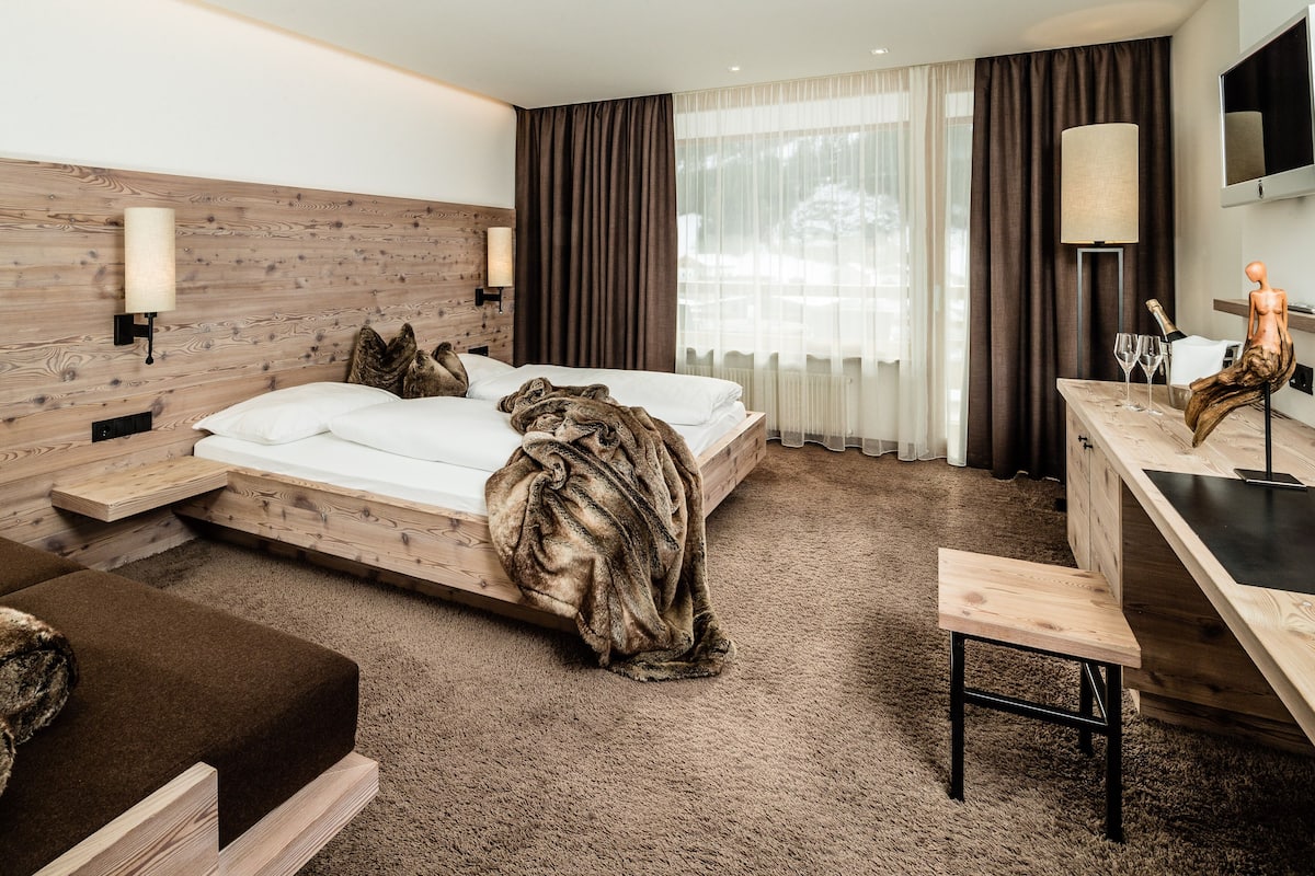 Ein modernes Hotelzimmer mit einem Holzkopfende, einem großen Bett, einem Holztisch, einer Bank und einem großen Fenster mit Vorhängen.