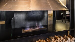 Une cheminée moderne dotée d’une hotte en métal, avec du bois de chauffage rangé en dessous et un feu visible qui brûle à l’intérieur.