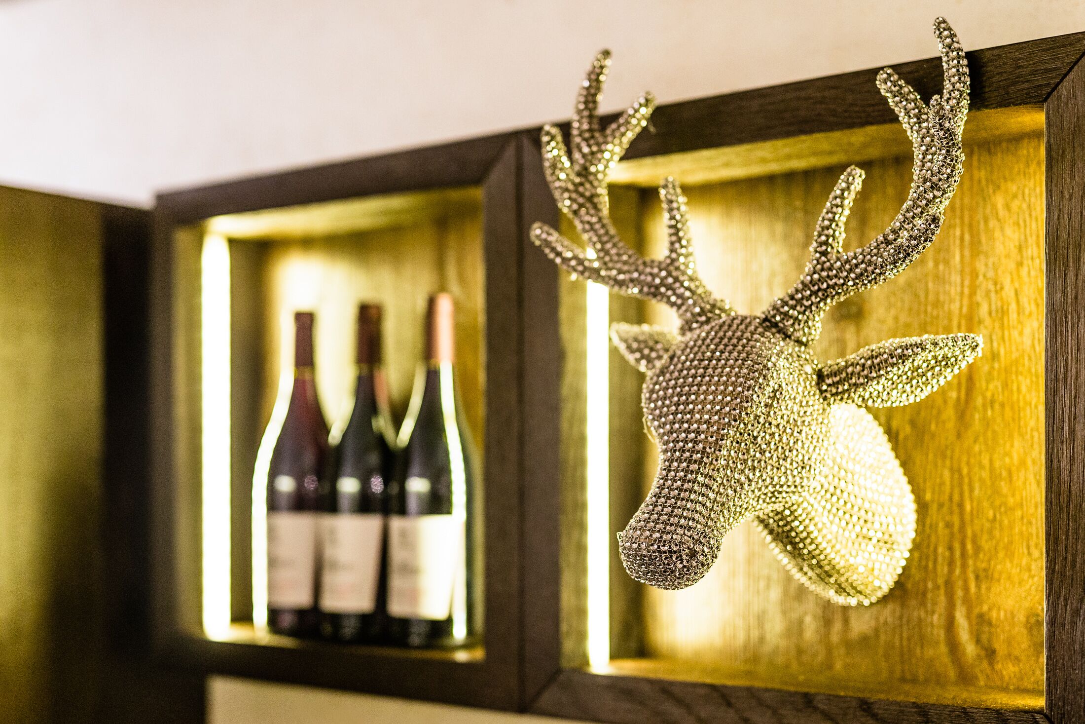 Een decoratief hertenbeeld met strass-steentjes in een houten lijst, met flessen wijn op de achtergrond.