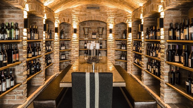 A wine cellar with stone walls, wooden shelves, and a central table with chairs. - Boutique & Fashion Hotel Maciaconi (Selva di Val Gardena)