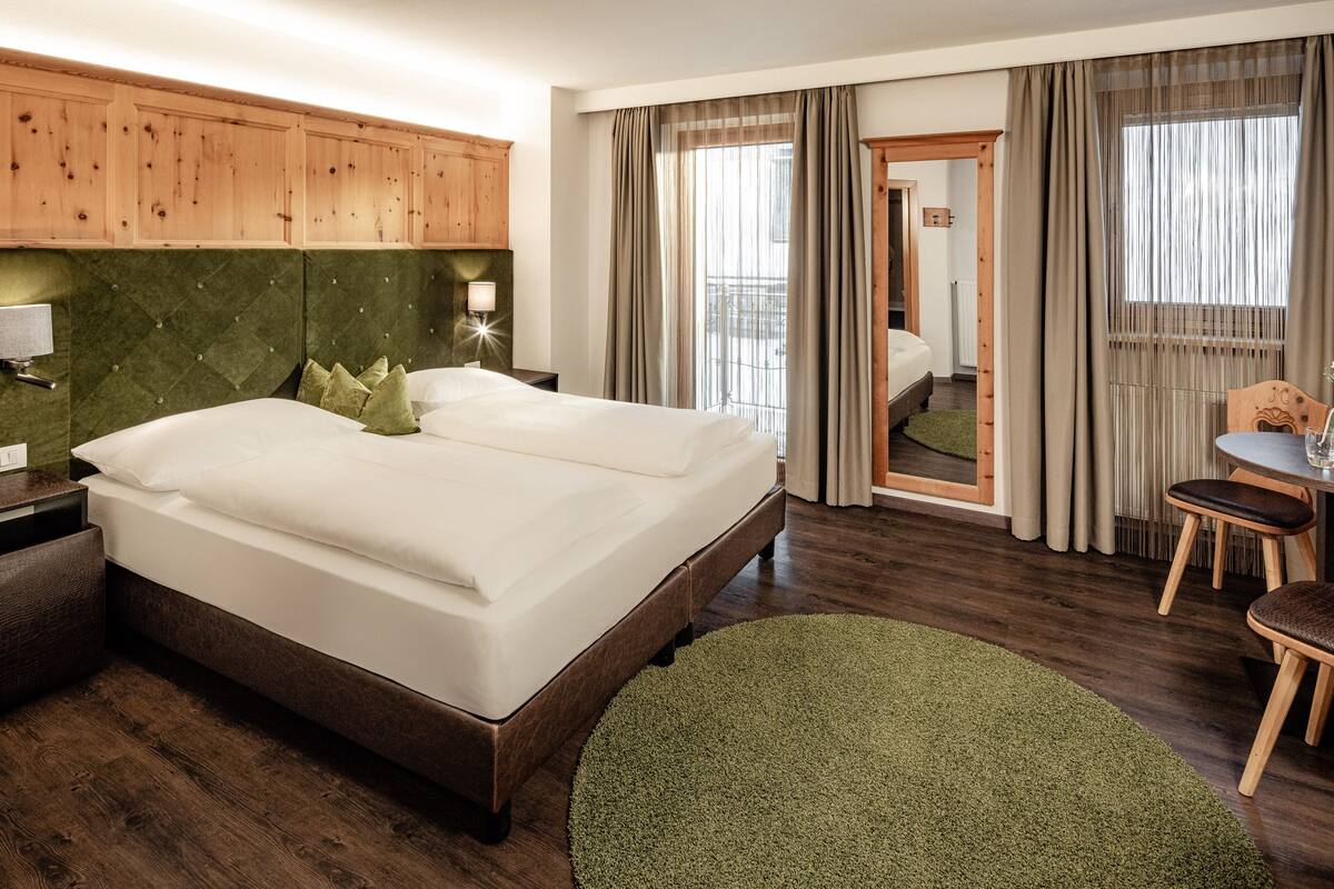 A hotel room with a large bed, wooden headboard, a small table with chairs, and a view of the balcony through the window.