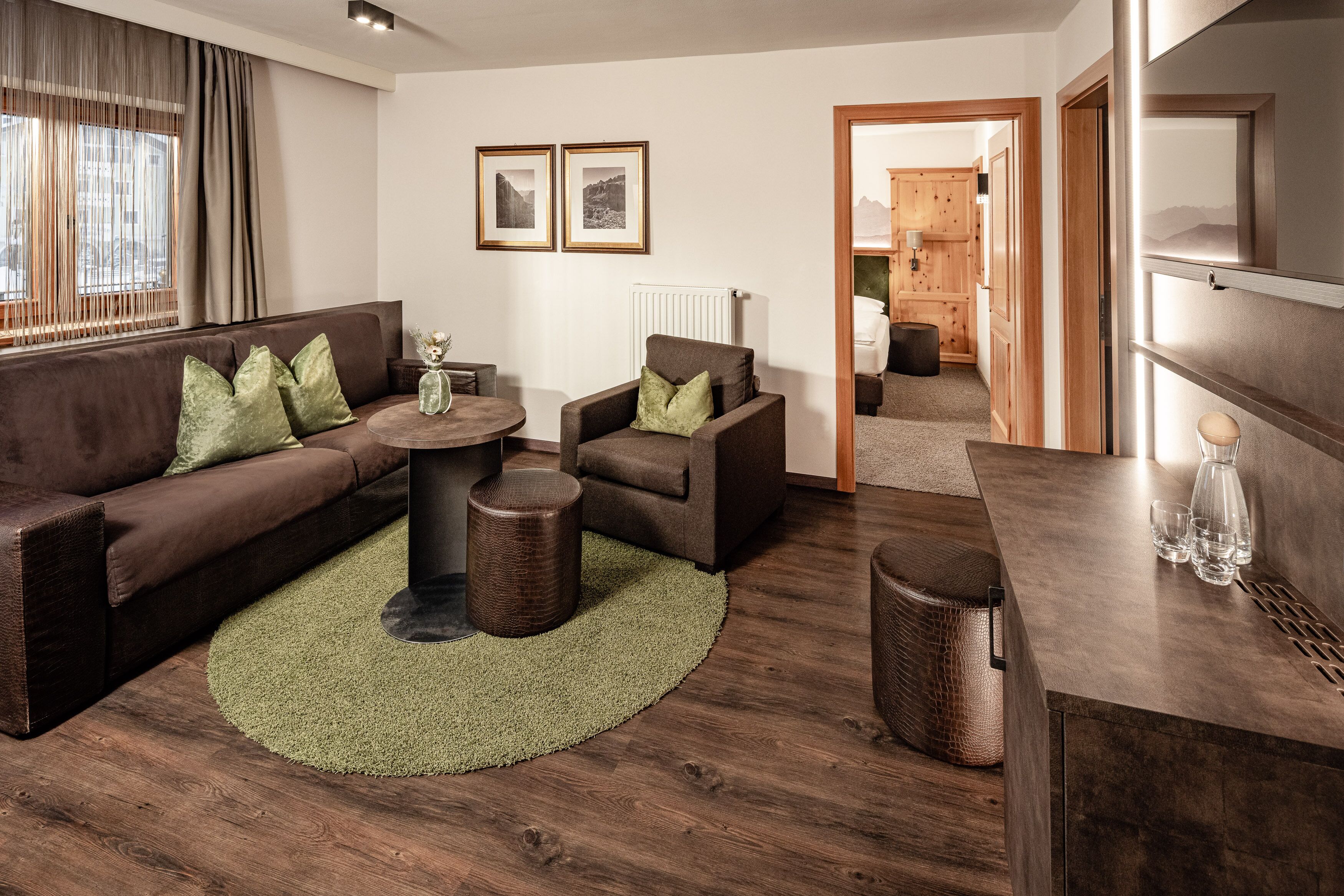 Una sala de estar moderna con un sofá marrón, dos sillones, una mesa de centro redonda y un tapiz verde. Hay cuadros enmarcados en la pared, un radiador y un aparador de madera con vasos.