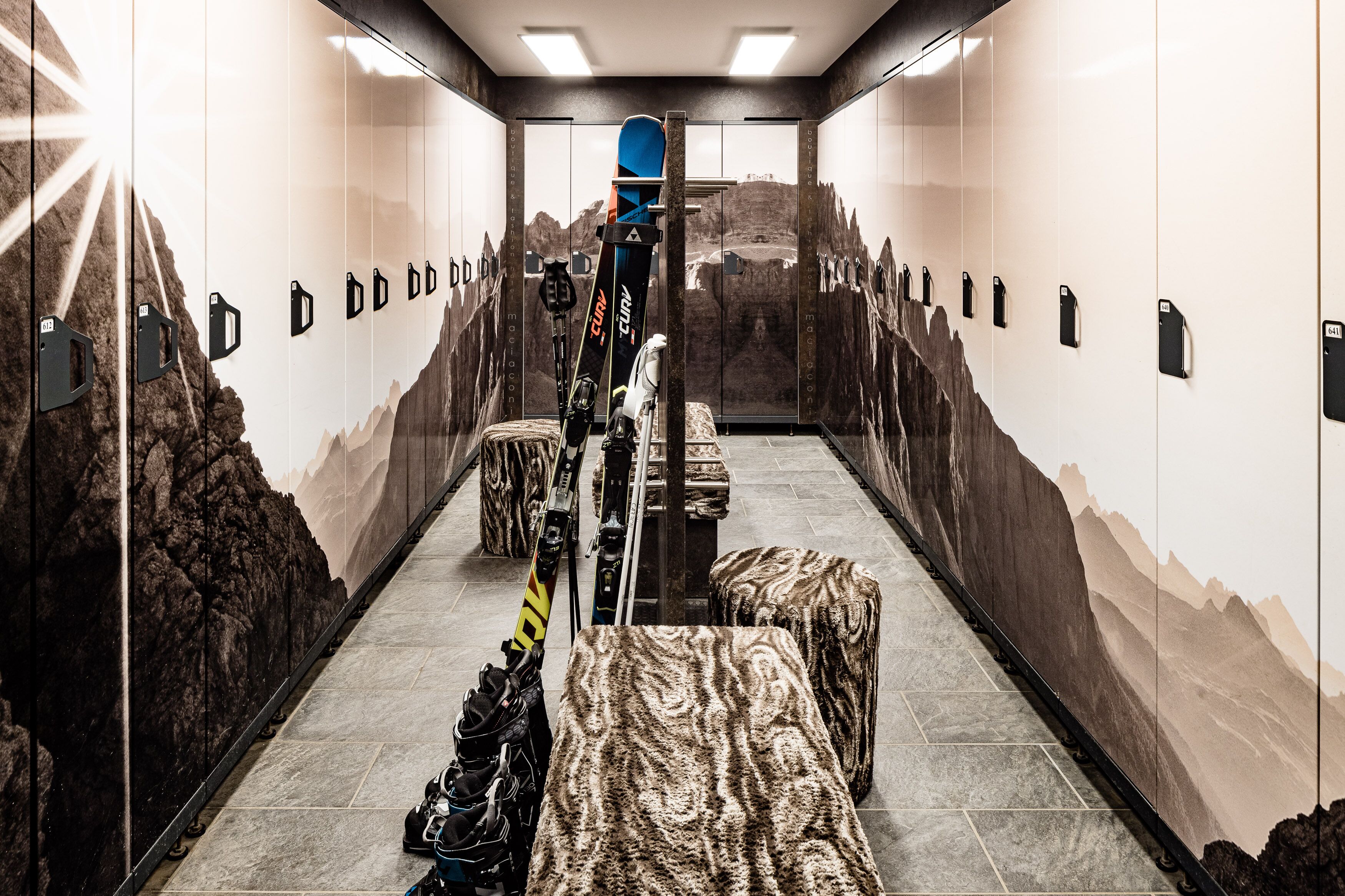 A ski lift area with a mountain mural on the walls, a ski lift chair, and a ski rack with skis and poles.