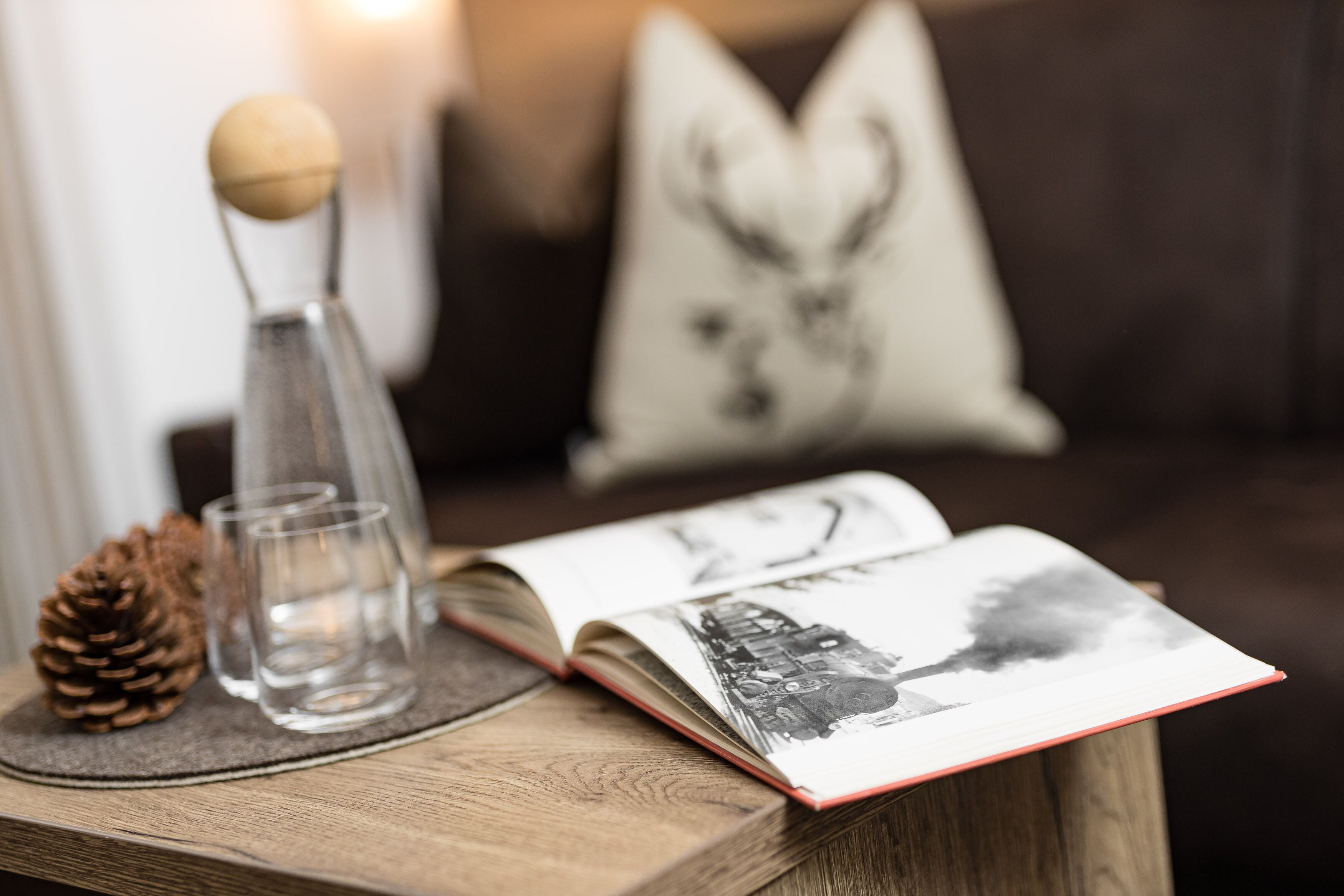 A book with a black and white image on the cover, a pine cone, and two clear glasses on a wooden table.