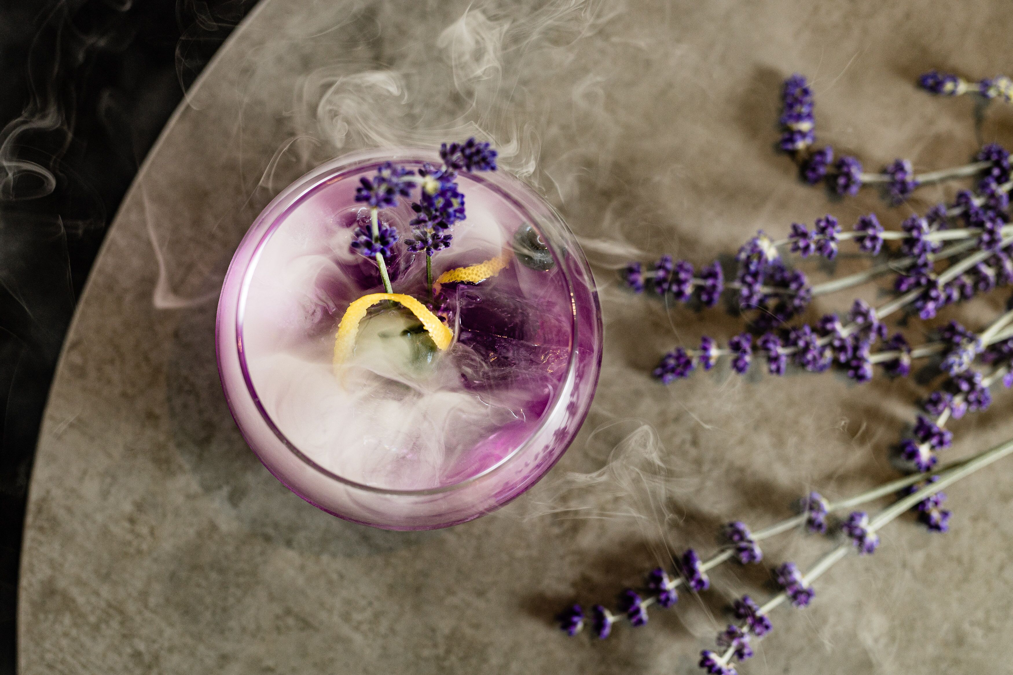 A purple glass with a lemon slice and lavender, surrounded by smoke and lavender branches.