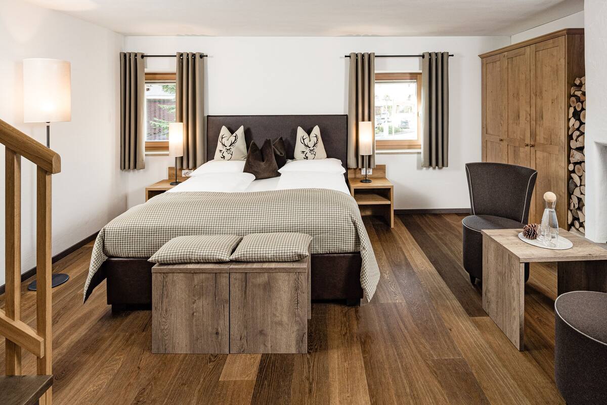 A modern hotel room with a large bed, wooden flooring, a seating area with a table and chairs, a fireplace, and a view of the outdoors.