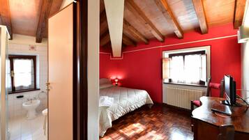 Quarto com teto de madeira, parede vermelha, cama, escrivaninha com computador e janela.