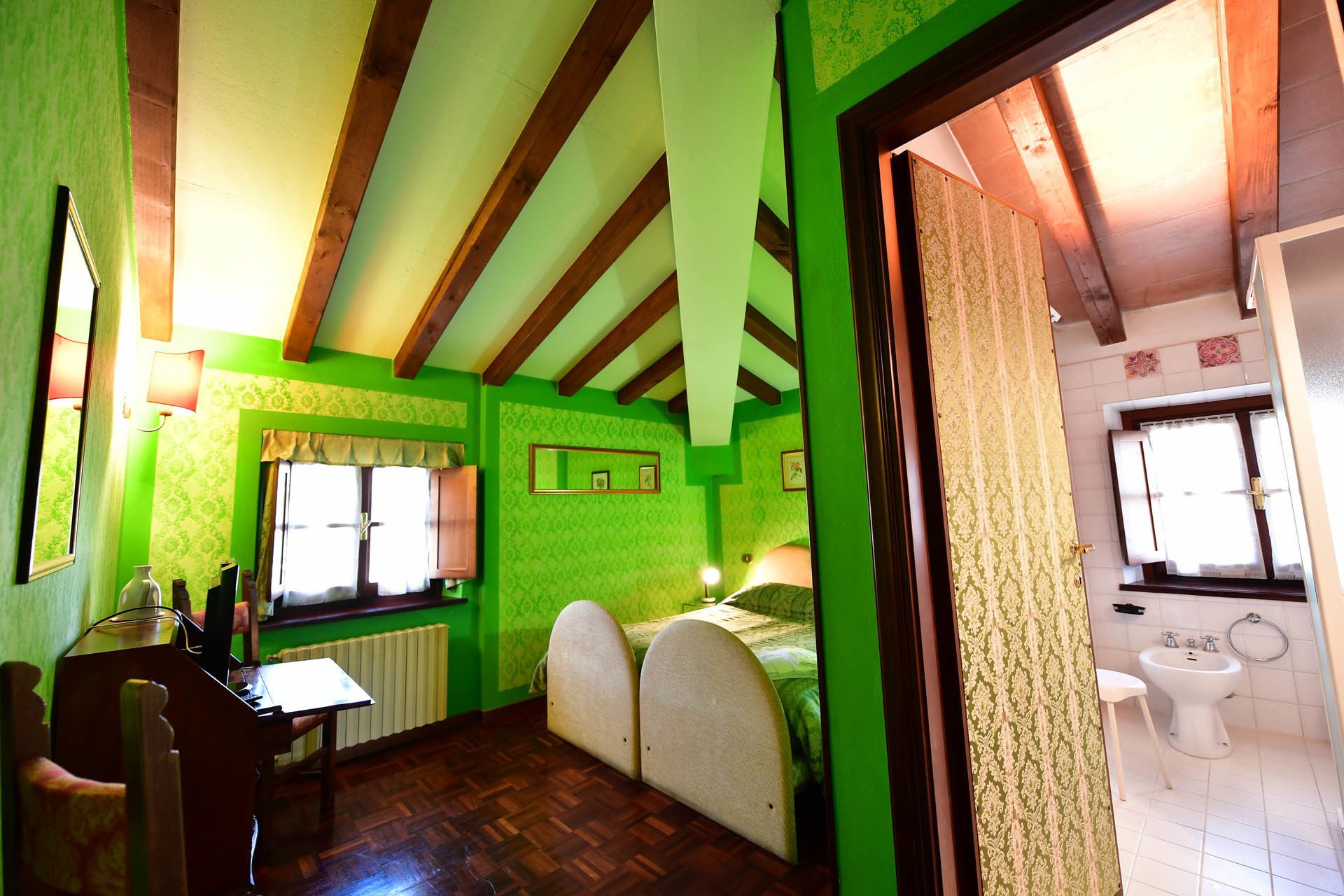 Quarto com teto de madeira, paredes verdes, cama com cabeceira, escrivaninha com cadeira, radiador e janela.