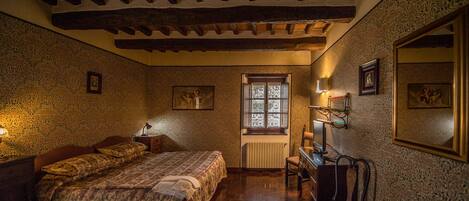 A bedroom with a wooden ceiling, patterned wallpaper, a bed with a patterned bedspread, a chair, a desk, and a window with a radiator below.