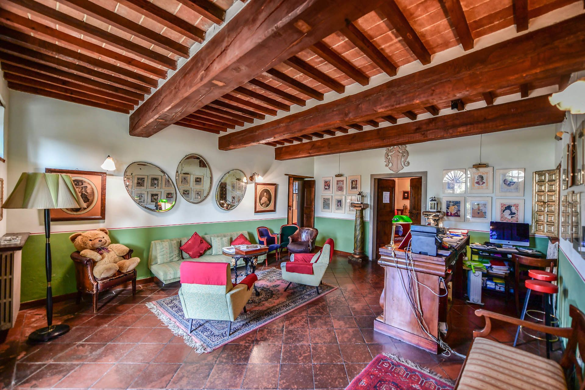 A spacious living room with a high wooden ceiling, green walls, and a tiled floor. There are several chairs, a sofa, and a table with various items on it.