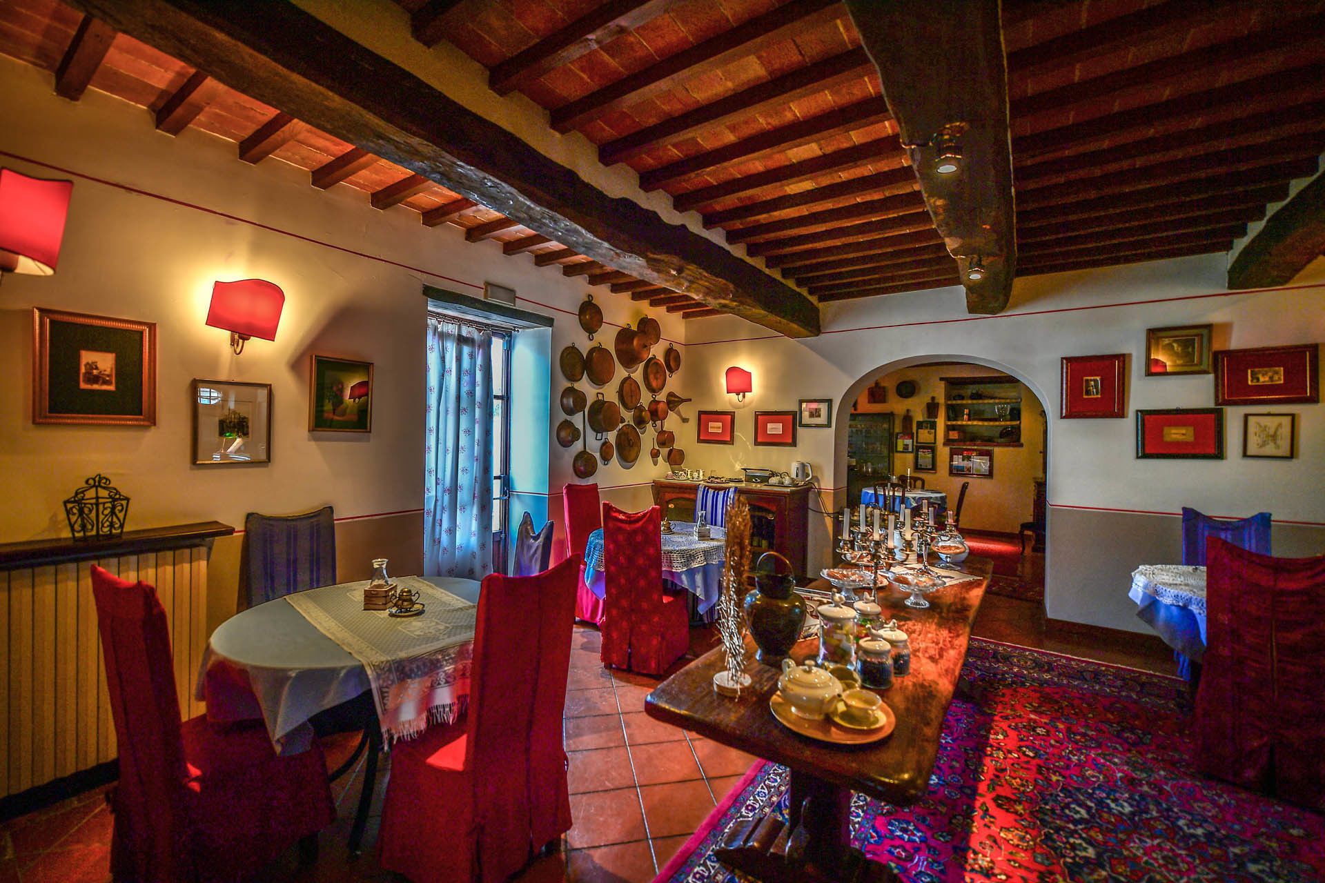 A dining area with a wooden ceiling, red chairs, a central table with food and drink, and a patterned rug.