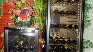 Een wijnkoeler met meerdere planken vol wijnflessen, een mand met decoratief groen en een rode houten lattenstructuur op de achtergrond.