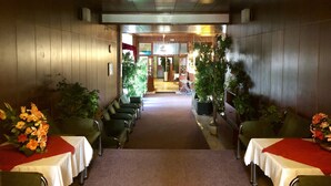 Een hotelgang met een tapijtvloer, groen gestoffeerde stoelen en tafels met witte tafelkleden. Aan weerszijden van de gang staan potplanten.