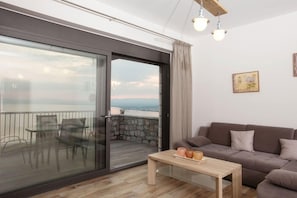 A modern living room with a glass sliding door leading to a balcony, a brown sofa, a wooden coffee table, and a painting on the wall. - Petra Thea (Kalamata)