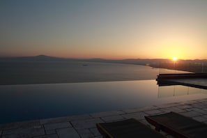 An infinity pool with a view of the sunset over the sea, a lounge area with chairs, and a clear sky. - Petra Thea (Kalamata)