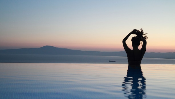 A person standing in a reflective pool with a view of distant mountains and a ship in the water. - Petra Thea (Kalamata)