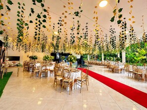 A spacious indoor venue with multiple round tables and chairs, a red carpet, and hanging foliage decorations.