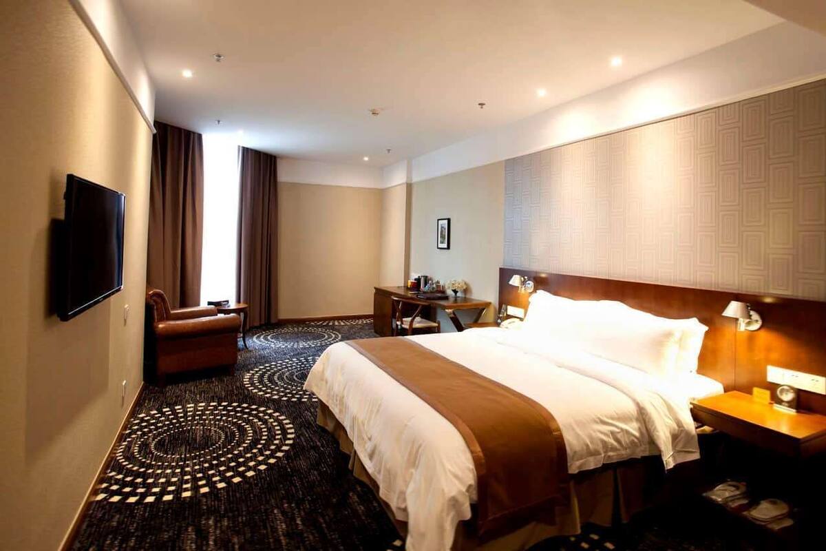 A hotel room with a large bed, a desk, a chair, a television, and a patterned carpet.