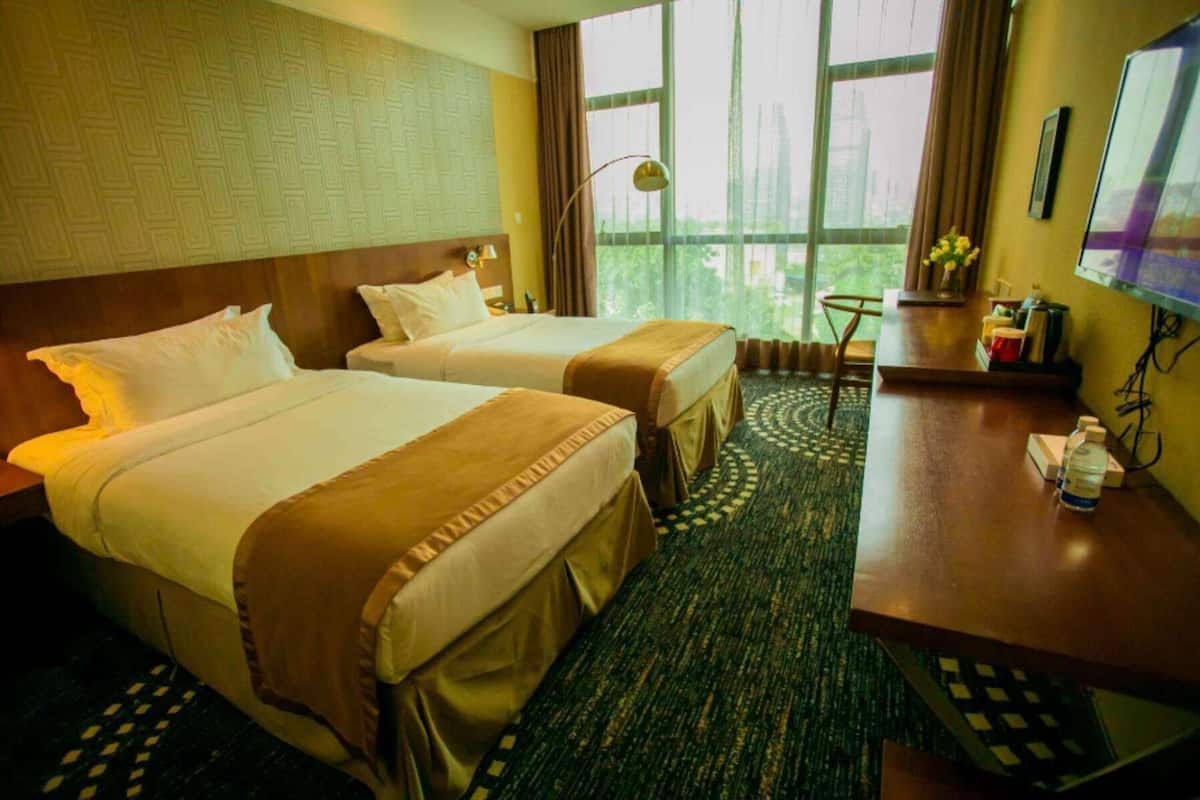 A hotel room with two beds, a desk, a chair, a lamp, and a view of the city through large windows.