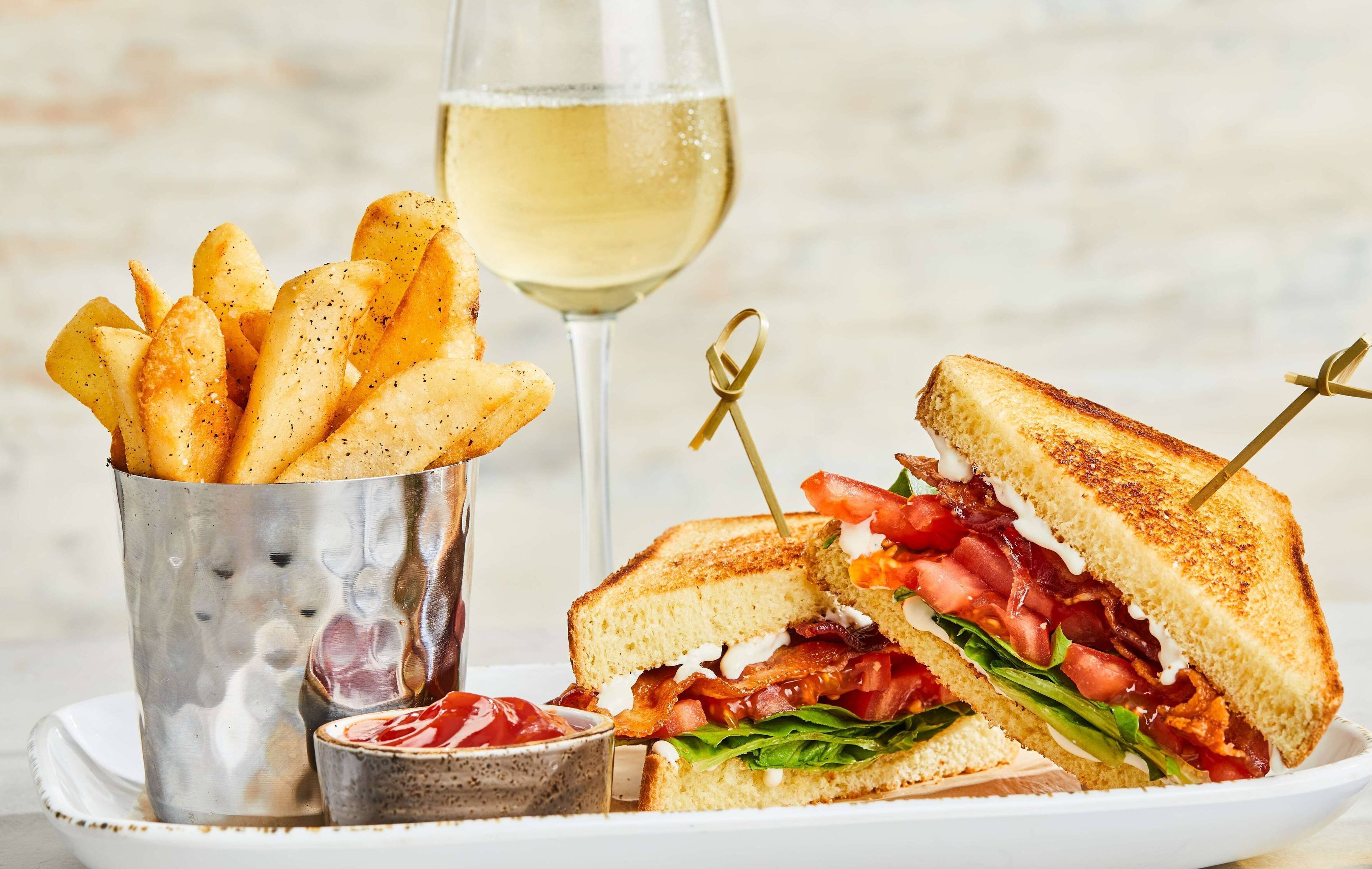A plate with a grilled sandwich, a glass of white wine, and a container of fries.