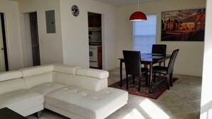 Una sala de estar con un sofá seccional blanco, una mesa de comedor con cuatro sillas, una lámpara colgante roja y un cuadro de un paisaje urbano.