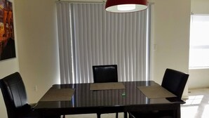 Un comedor con una mesa rectangular, cuatro sillas, una lámpara colgante roja y persianas verticales.