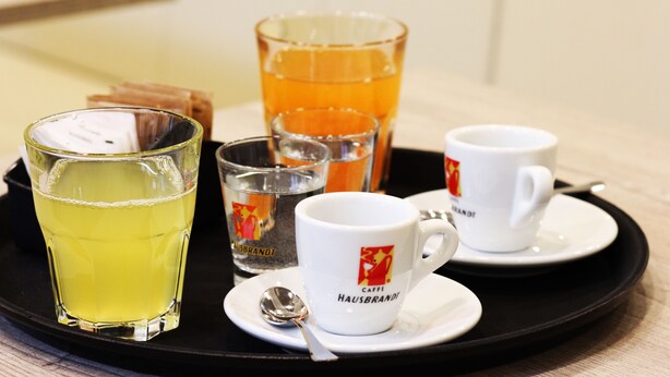 A tray with a glass of yellow liquid, a glass of orange liquid, and two white cups with saucers and a spoon. - Sole Hotel Verona (Verona)