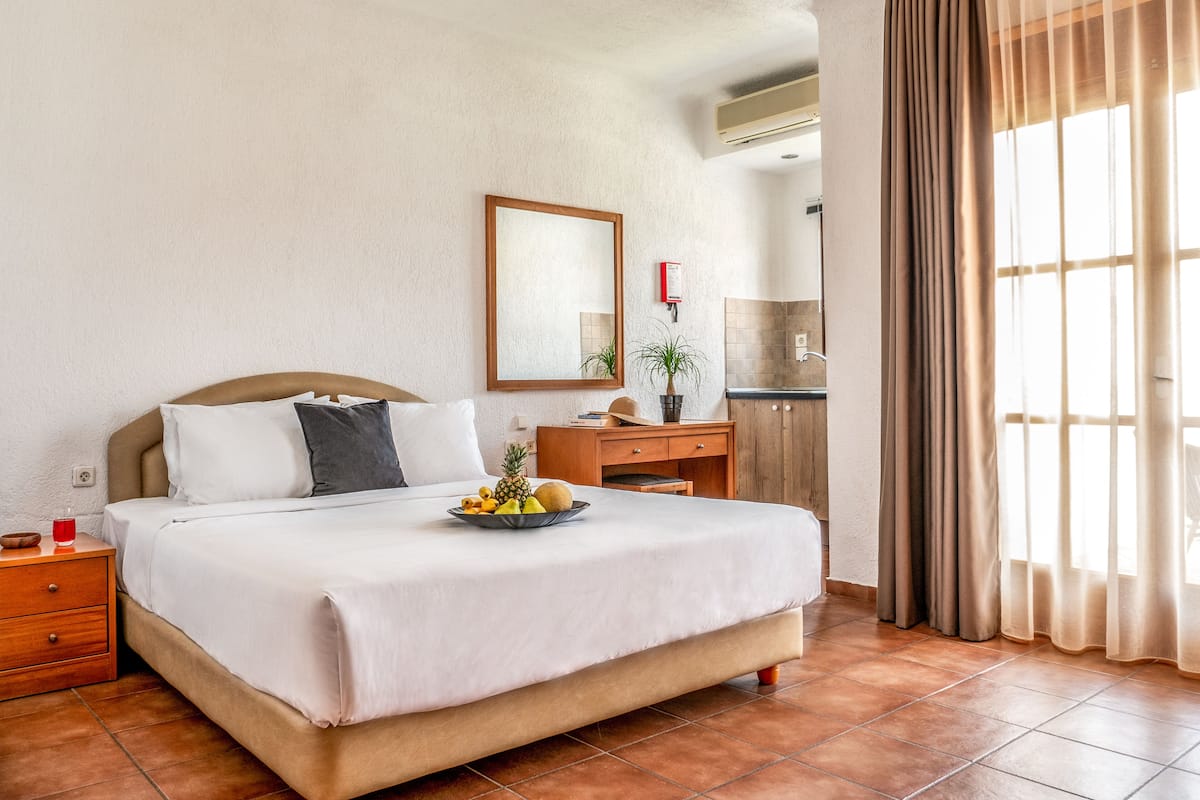 A hotel room with a bed, a nightstand, a dresser, a mirror, a small table with fruit, and a window with curtains.