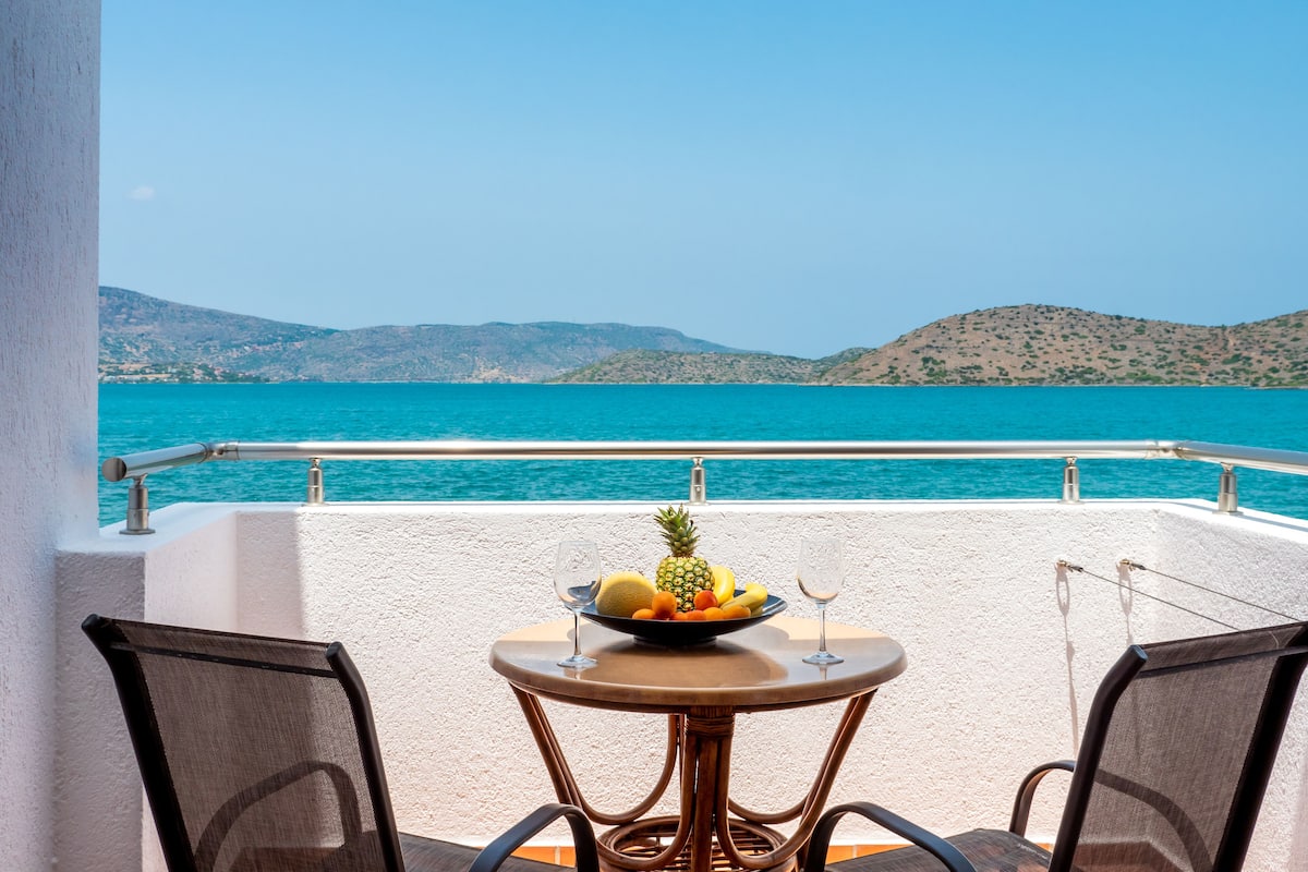 A balcony with a table set for two, overlooking a turquoise sea and distant hills.