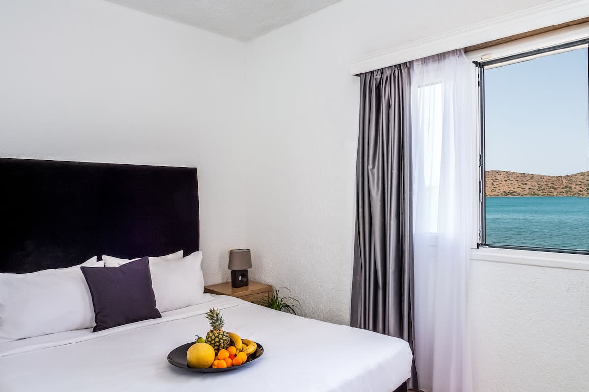A hotel room with a bed, a bedside table with a lamp, a fruit plate on the bed, and a view of the sea through a window.