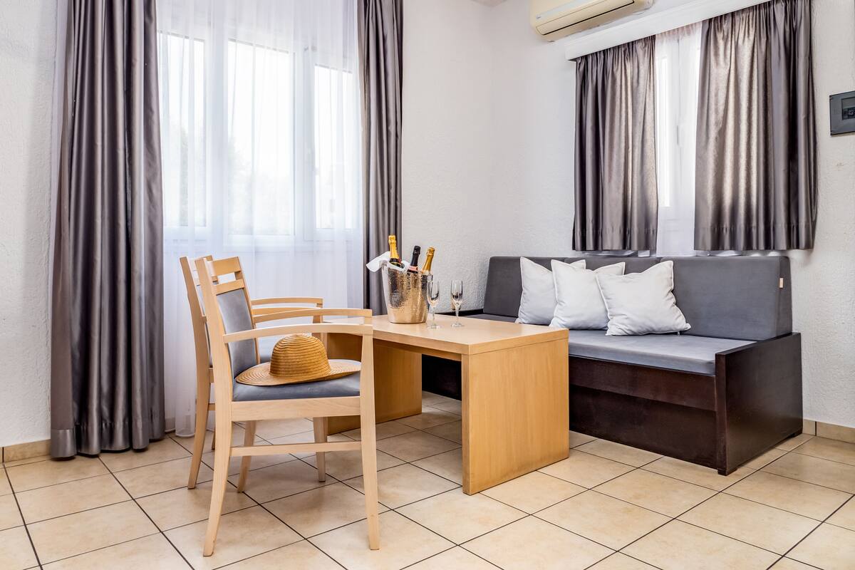 A modern room with a wooden desk, a grey sofa, and a chair. There is a window with curtains, an air conditioning unit, and a tiled floor.