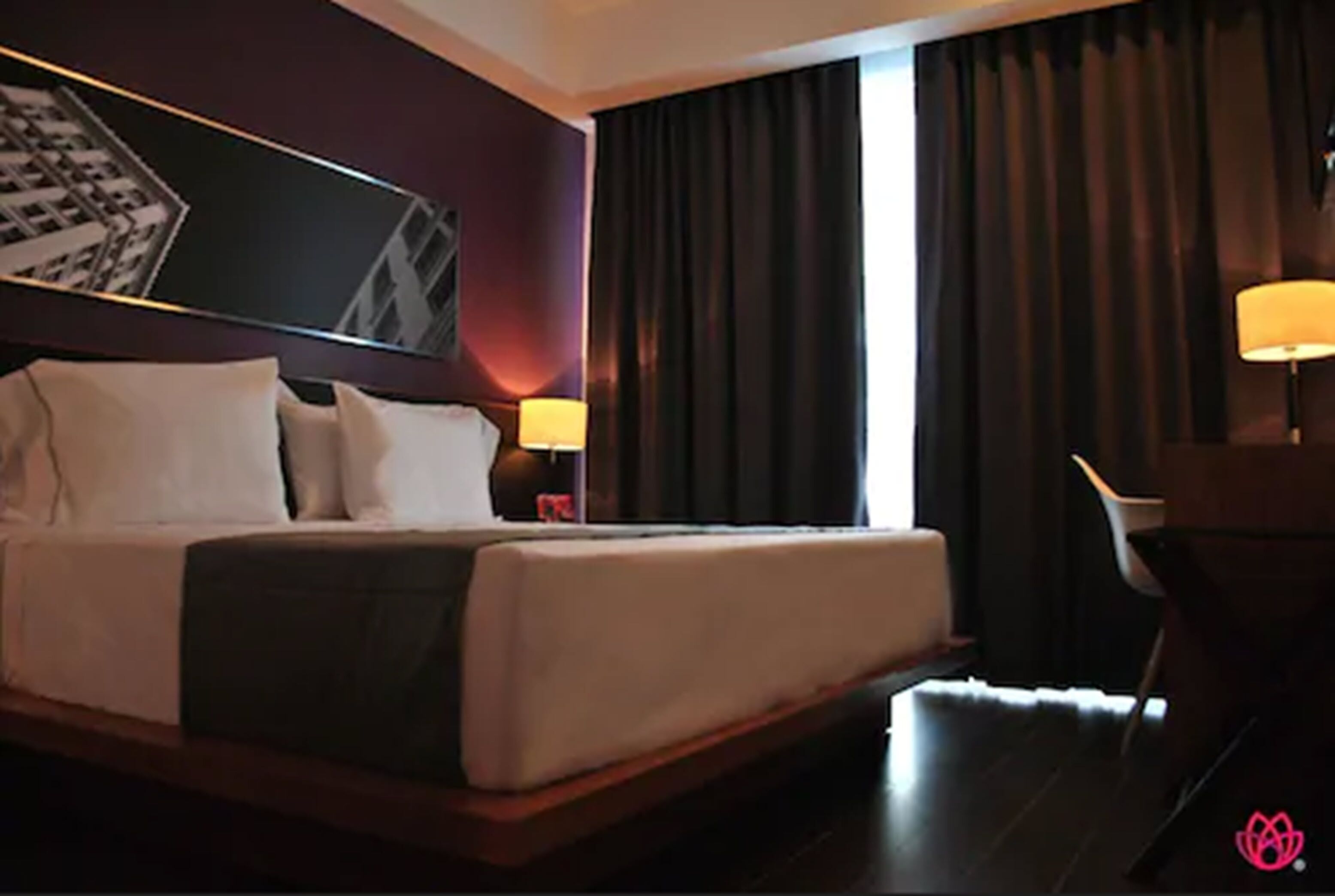 Una camera d'albergo con un letto grande, un comodino con una lampada, una sedia e vista su un edificio dalla finestra.