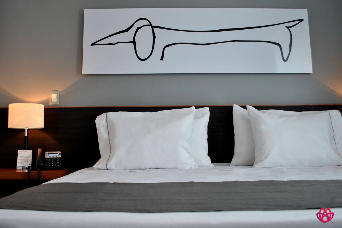 A hotel room with a bed, white pillows, a grey blanket, a bedside lamp, a phone, and a wall-mounted artwork.