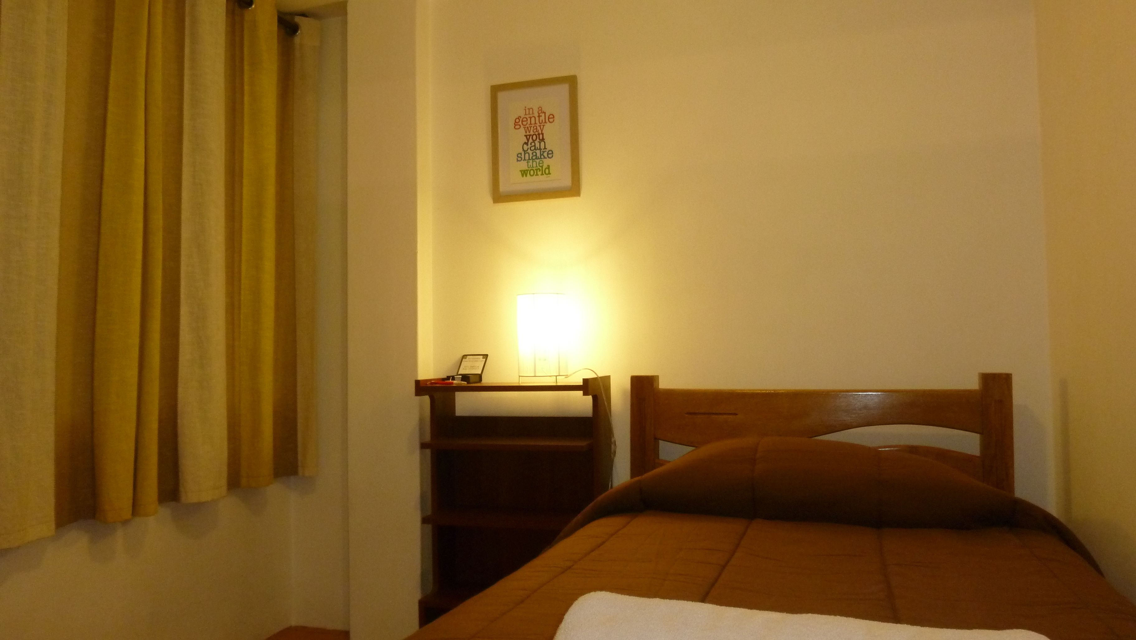 Una cama individual con cabecera de madera, una mesita de noche con lámpara, y una cita inspiradora enmarcada en la pared.