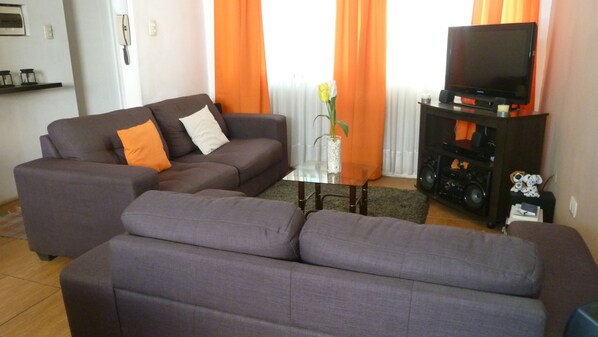 A living room with a sectional sofa, a glass coffee table, a flat-screen TV, and a small table with speakers and decorative items.