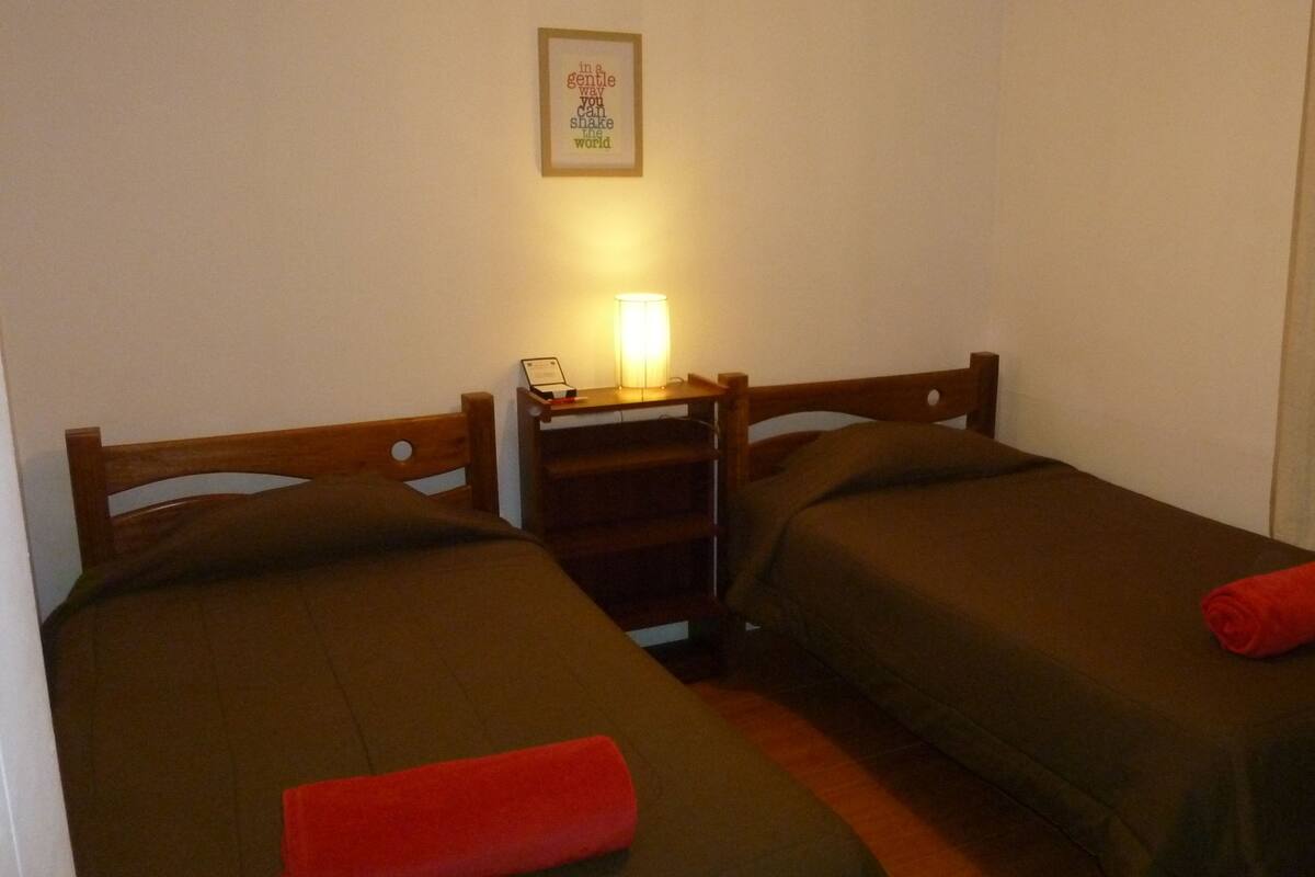 A small, dimly lit room with two single beds, a nightstand with a lamp, and a framed inspirational quote on the wall.