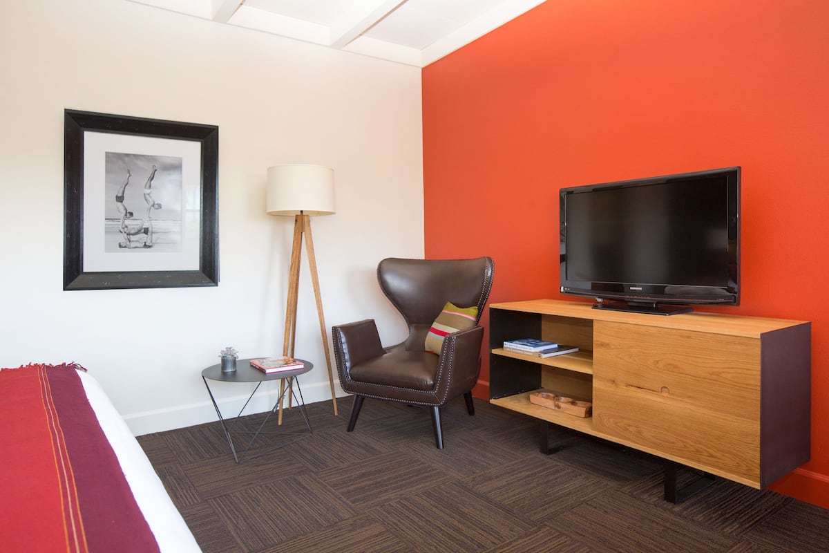 A hotel room with a bed, a chair, a TV on a wooden stand, a framed picture on the wall, and a floor lamp.
