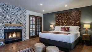 A bedroom with a fireplace, a bed with pillows, two ottomans, a striped rug, and a wall with a geometric pattern.